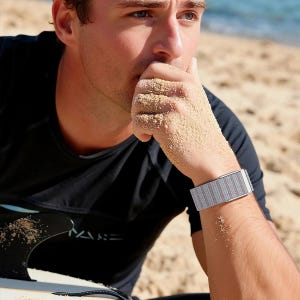 May include: A watch band featuring a woven fabric texture with alternating vertical stripes in light gray and muted reddish-brown tones, secured with a metallic silver buckle clasp, worn on a wrist covered in fine sand particles.