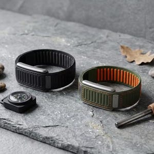 May include: Two silicone watch bands, one solid black and one olive green with a bright orange interior lining, featuring vertical ribbed textures and metallic silver hardware, accompanied by a small black compass and a metal fire starter rod.