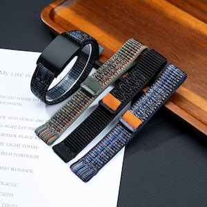 May include: Four watch bands made of woven nylon fabric. One band is looped with a black metal connector, while the other three are flat. The colors include a dark blue, a green and orange mix, a solid black with an orange loop, and a blue and orange pattern.