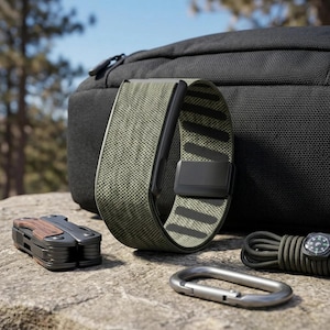 May include: A smartwatch band featuring a textured olive green fabric exterior with a black interior lining and a black magnetic clasp. The band is positioned upright, showcasing its flexible construction and the contrast between the green weave and black hardware.