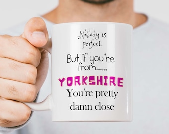 Può includere: Una tazza in ceramica bianca con un design nero e rosa. Il testo sulla tazza recita "Nobody is perfect. But if you're from...... YORKSHIRE You're pretty damn close."