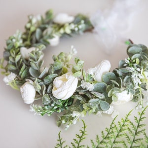 May include: A white flower crown with green leaves and white flowers. The crown is made of artificial flowers and is perfect for a wedding or other special occasion.