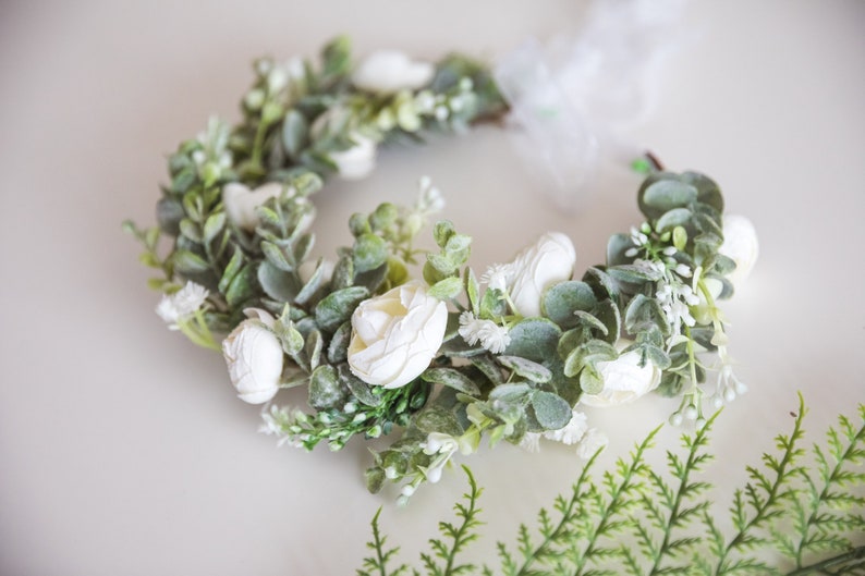 May include: A white flower crown with green leaves and white flowers. The crown is made of artificial flowers and is perfect for a wedding or other special occasion.