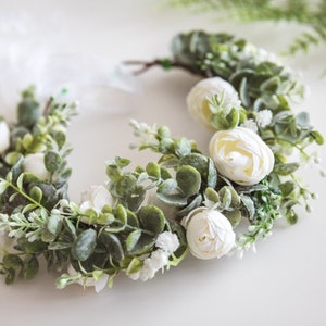 May include: A white floral crown with white flowers and green leaves. The crown is made of artificial flowers and is perfect for a wedding or other special occasion.