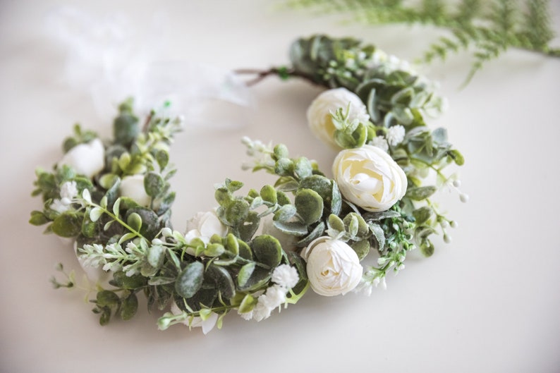 May include: A white floral crown with white flowers and green leaves. The crown is made of artificial flowers and is perfect for a wedding or other special occasion.