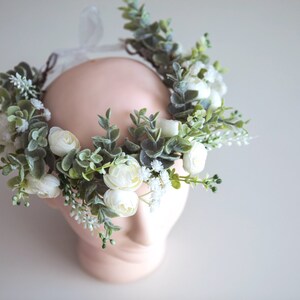 May include: A white flower crown with green leaves and white flowers. The crown is made of artificial flowers and is perfect for a wedding or other special occasion.