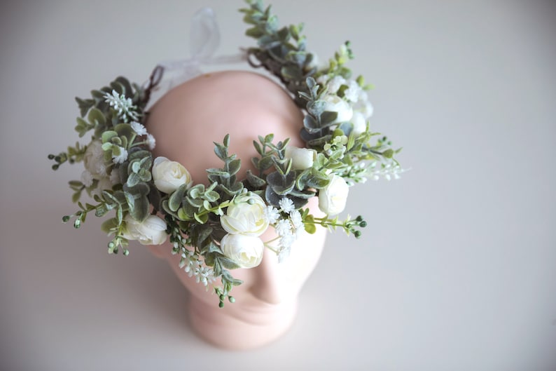 May include: A white flower crown with green leaves and white flowers. The crown is made of artificial flowers and is perfect for a wedding or other special occasion.