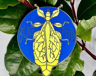 Puede incluir: Un pin redondo de esmalte azul y amarillo con un diseño de insecto estilizado. El pin presenta las palabras "BUG FAN" en letras negras. El insecto es amarillo con contornos negros. El pin está sobre un fondo de hojas verdes.
