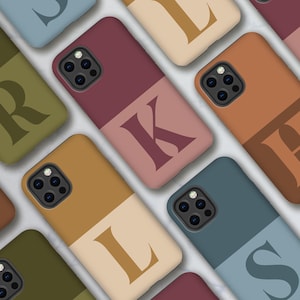 Puede incluir: Una colección de fundas de teléfono coloridas, cada una con un diseño de dos tonos y una letra grande. Las fundas son en tonos burdeos, verde oliva, beige y azul, con letras en colores complementarios. Disposición diagonal.