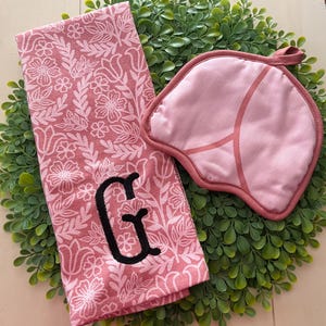 May include: A pink floral patterned kitchen towel with a black embroidered "G" and a matching pink pot holder. The towel and pot holder are on a green wreath.