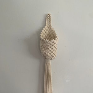May include: A white macrame wall hanging with a small pouch and a long tassel. The hanging is made of natural cotton rope and is perfect for adding a touch of bohemian style to any room.