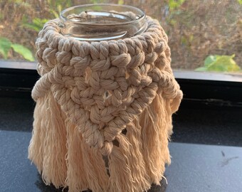 May include: A beige macrame candle holder with a glass jar inside and fringe detailing.
