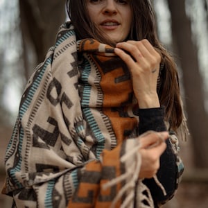 Könnte beinhalten: Eine Frau trägt einen braunen, schwarzen und blauen gemusterten Wollschal mit Fransen. Der Schal ist um ihren Hals und ihre Schultern gewickelt.