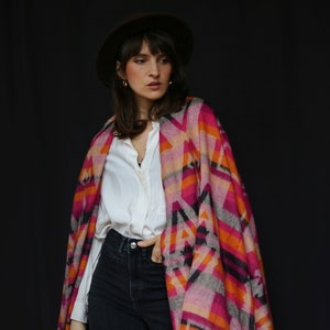 Könnte beinhalten: Ein farbenfroher Schal mit geometrischem Muster in Rosa, Orange und Schwarz. Der Schal hat einen Fransenrand und wird über einem weißen Hemd und dunklen Jeans getragen. Ein brauner Hut rundet den Look ab.