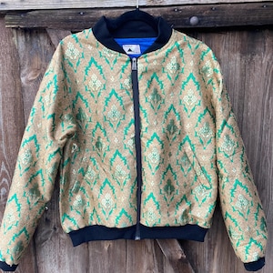 Könnte beinhalten: Eine Bomberjacke mit gold-grünem Damastmuster, schwarzem Rippstrickkragen, Bündchen und Saum. Die Jacke hat einen schwarzen Reißverschluss in der Mitte und ein blaues Innenfutter. Ein weißes Etikett mit schwarzem Text ist am Halsausschnitt sichtbar.