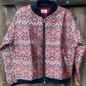 Könnte beinhalten: Eine Bomberjacke mit leuchtend roter Grundfarbe und einem komplexen, sich wiederholenden Muster in Gold, Türkis und Violett. Die Jacke hat einen schwarzen Rippstrickkragen, Bündchen, einen Saum, einen Frontreißverschluss und ein AODA-Etikett.