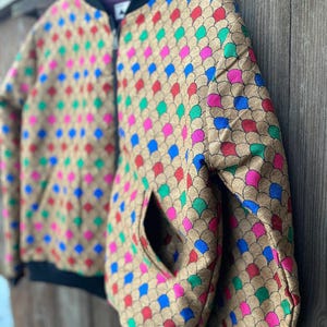 Könnte beinhalten: Eine Bomberjacke mit beigefarbener Basis und einem sich wiederholenden Muster aus überlappenden Schuppen in leuchtenden Blau-, Rot-, Grün- und Rosatönen. Die Jacke hat einen schwarzen Rippstrickkragen, schwarze Rippstrickbündchen und einen schwarzen Reißverschluss.