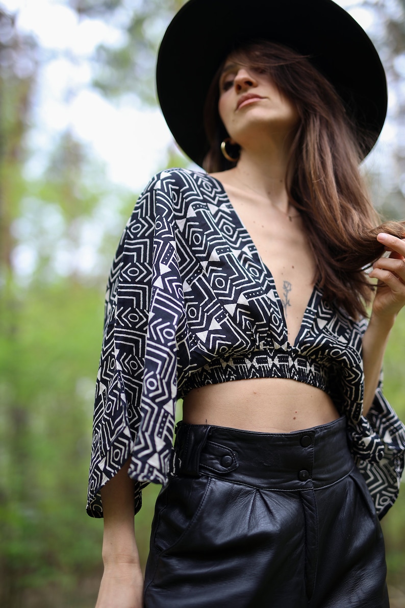 Op de afbeelding: Een vrouw draagt een zwarte hoed met brede rand, een zwart-wit geometrisch geprint crop top en een zwarte leren broek.
