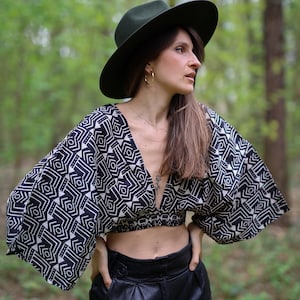 Op de afbeelding: Een vrouw draagt een zwart-wit geometrisch geprint crop top met een diepe V-hals en wijde mouwen. Ze draagt ook een zwarte leren broek en een groene fedora.