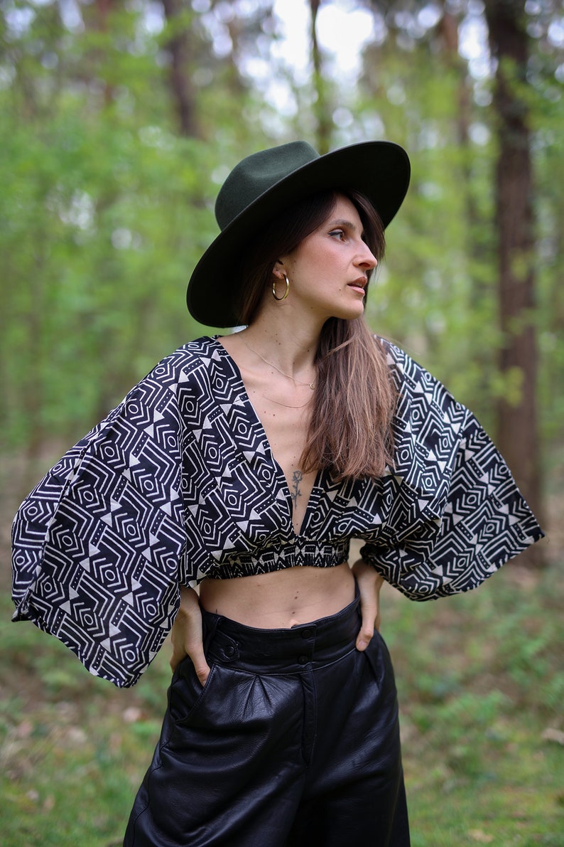Op de afbeelding: Een vrouw draagt een zwart-wit geometrisch geprint crop top met een diepe V-hals en wijde mouwen. Ze draagt ook een zwarte leren broek en een groene fedora.