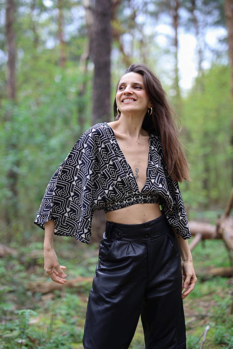 Op de afbeelding: Een vrouw draagt een zwart-wit geometrisch geprint crop top met een diepe V-hals en lange mouwen. Ze draagt ook een zwarte leren broek met een hoge taille.