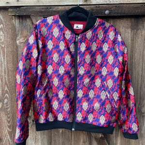 Könnte beinhalten: Eine Bomberjacke mit einem leuchtend blauen und roten, gesprenkelten Hintergrund und einem sich wiederholenden, goldfarbenen blattförmigen Muster. Die Jacke hat einen schwarzen Rippstrickkragen, Bündchen, einen Saum, einen Reißverschluss und ein Etikett mit der Aufschrift ADREN STUDIOS.