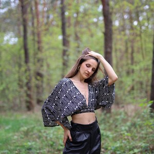 Op de afbeelding: Een vrouw draagt een zwart-wit geometrisch geprint crop top met een zwarte leren broek in een bosrijke omgeving.