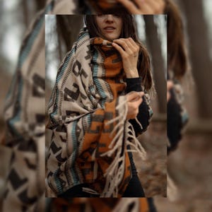 May include: A large, rectangular woven blanket wrap featuring a geometric pattern in shades of cream, burnt orange, dark brown, and teal. The fabric has a textured appearance with long, cream-colored fringe tassels along the bottom edge.