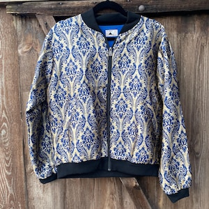 May include: A bomber jacket featuring a gold and blue damask pattern. The jacket has a black ribbed collar, black ribbed cuffs, and a black ribbed hem. A central black zipper runs down the front, and a blue interior lining is visible at the neckline.