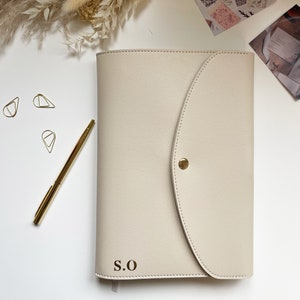 May include: A cream leather planner with a flap closure and a gold button. The planner has the initials "S.O." embossed on the front.