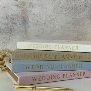 Peut inclure: Quatre carnets de planification de mariage empil&eacute;s les uns sur les autres. Les carnets sont de couleurs diff&eacute;rentes&nbsp;: blanc, beige, bleu clair et rose. Chaque carnet porte l&rsquo;inscription &laquo;&nbsp;Wedding Planner&nbsp;&raquo; en or sur la couverture.