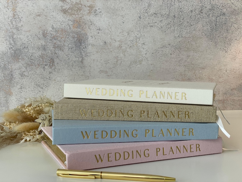 Peut inclure: Quatre carnets de planification de mariage empil&eacute;s les uns sur les autres. Les carnets sont de couleurs diff&eacute;rentes&nbsp;: blanc, beige, bleu clair et rose. Chaque carnet porte l&rsquo;inscription &laquo;&nbsp;Wedding Planner&nbsp;&raquo; en or sur la couverture.