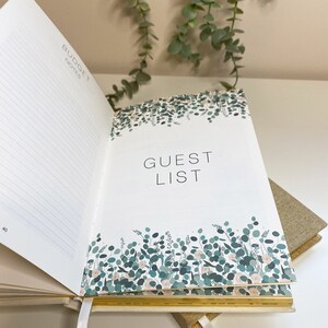 Peut inclure: Un planificateur de mariage ouvert avec un fond blanc et des accents dor&eacute;s. La page de gauche porte l'en-t&ecirc;te "Budget Notes" et du papier lign&eacute;. La page de droite porte l'en-t&ecirc;te "Guest List" avec un motif floral de feuilles vertes et de fleurs blanches.