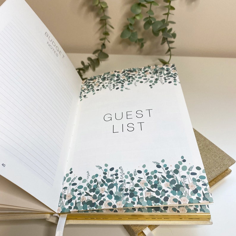 Peut inclure: Un planificateur de mariage ouvert avec un fond blanc et des accents dor&eacute;s. La page de gauche porte l'en-t&ecirc;te "Budget Notes" et du papier lign&eacute;. La page de droite porte l'en-t&ecirc;te "Guest List" avec un motif floral de feuilles vertes et de fleurs blanches.