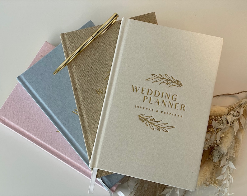 Peut inclure: Une pile de quatre journaux de planification de mariage avec un stylo dor&eacute; sur le dessus. Le journal du dessus est de couleur cr&egrave;me avec le texte "Wedding Planner Journal & Keepsake" en lettres dor&eacute;es. Les autres journaux sont rose, bleu et marron.