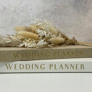 Peut inclure: Deux livres de planification de mariage reli&eacute;s en lin blanc empil&eacute;s l'un sur l'autre. Le livre du dessus a une couverture en lin beige avec le texte "Wedding Planner" en lettres dor&eacute;es. Le livre du dessous a une couverture en lin blanc avec le texte "Wedding Planner" en lettres dor&eacute;es. Un bouquet de fleurs s&eacute;ch&eacute;es blanches et beiges est pos&eacute; sur les livres.
