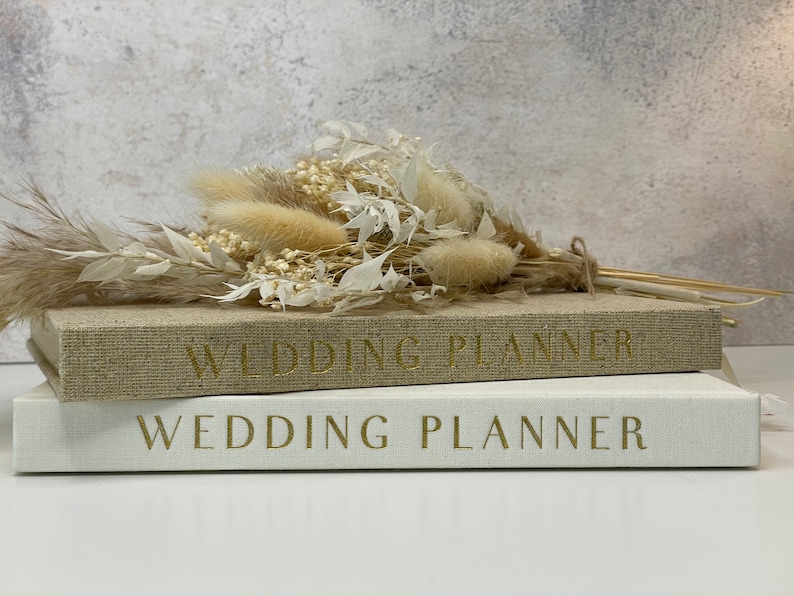 Peut inclure: Deux livres de planification de mariage reli&eacute;s en lin blanc empil&eacute;s l'un sur l'autre. Le livre du dessus a une couverture en lin beige avec le texte "Wedding Planner" en lettres dor&eacute;es. Le livre du dessous a une couverture en lin blanc avec le texte "Wedding Planner" en lettres dor&eacute;es. Un bouquet de fleurs s&eacute;ch&eacute;es blanches et beiges est pos&eacute; sur les livres.