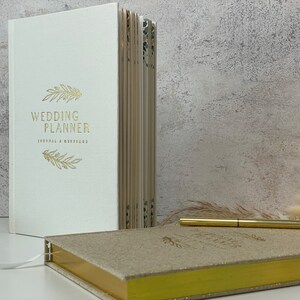 Peut inclure: Un carnet de planification de mariage blanc avec un lettrage dor&eacute; qui dit "Wedding Planner Journal & Workbook" et un motif floral dor&eacute;. Un deuxi&egrave;me carnet dor&eacute; plus petit avec une couverture scintillante est &eacute;galement visible.