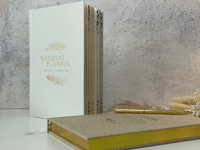 Peut inclure: Un carnet de planification de mariage blanc avec un lettrage dor&eacute; qui dit "Wedding Planner Journal & Workbook" et un motif floral dor&eacute;. Un deuxi&egrave;me carnet dor&eacute; plus petit avec une couverture scintillante est &eacute;galement visible.