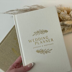 Peut inclure: Un carnet de planification de mariage blanc avec une inscription dor&eacute;e "Wedding Planner Journal & Keepsake" et un motif floral dor&eacute;.