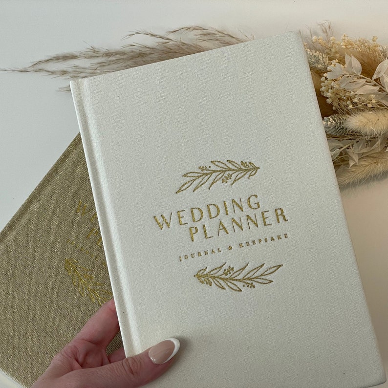Peut inclure: Un carnet de planification de mariage blanc avec une inscription dor&eacute;e "Wedding Planner Journal & Keepsake" et un motif floral dor&eacute;.