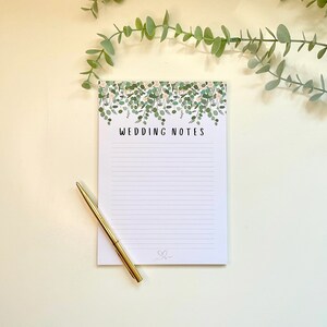 Puede incluir: Un bloc de notas blanco con un borde floral verde y el texto "Wedding Notes" en la parte superior. Un bolígrafo dorado está sobre el bloc de notas.