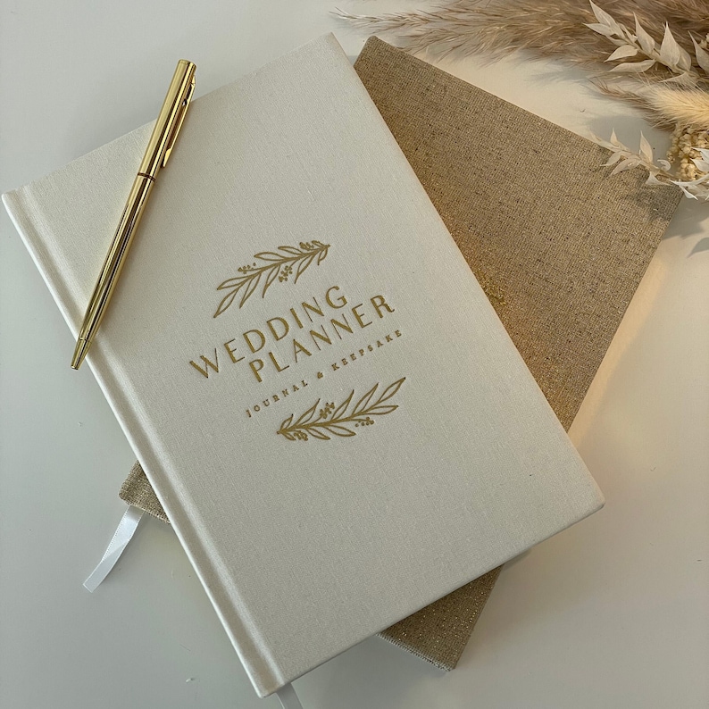 Peut inclure: Un carnet de planification de mariage blanc avec une inscription dor&eacute;e "Wedding Planner Journal & Keepsake" et un motif floral dor&eacute;.