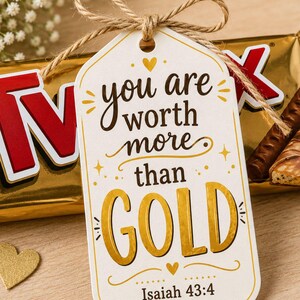 Può includere: Un'etichetta regalo con il messaggio "you are worth more than GOLD" in lettere dorate, legata con spago. L'etichetta è abbinata a una barretta di cioccolato avvolta in oro. Il versetto biblico Isaia 43:4 è stampato in basso.