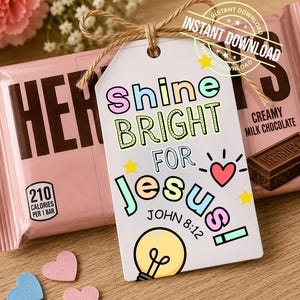 May include: A white rectangular gift tag with a jute twine bow, featuring the colorful text SHINE BRIGHT FOR JESUS! JOHN 8:12, a yellow lightbulb illustration, a red heart, and small yellow stars, placed in front of a pink Hershey's chocolate bar.
