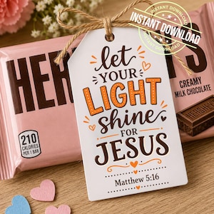 May include: A white tag with the text "let your light shine for Jesus" and "Matthew 5:16" is tied with twine to a pink Hershey's chocolate bar. Two pink and one blue heart-shaped confetti pieces are scattered near the tag.