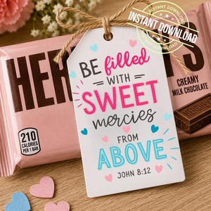Può includere: Un'etichetta regalo rettangolare bianca con un fiocco in corda di juta, recante il testo BE filled WITH SWEET mercies FROM ABOVE JOHN 8:12 in rosa, blu e nero, decorata con piccoli cuori rosa e blu, posizionata su una barretta di cioccolato Hershey's.