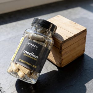 May include: A clear glass bottle filled with beige capsules, labeled "medina" by Jahn Herbs. The bottle has a black cap and a black and yellow label. A small wooden box sits behind the bottle on a dark surface.