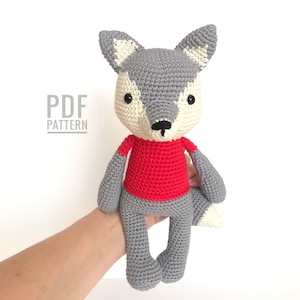 May include: A grey crocheted wolf wearing a red shirt. The wolf has white accents on its ears, nose, and tail. The wolf is sitting on a hand.
