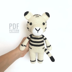 May include: A crocheted tiger stuffed animal with black and white stripes. The tiger has a white face with black eyes and a black nose. The tiger is standing on a white background. PDF PATTERN.